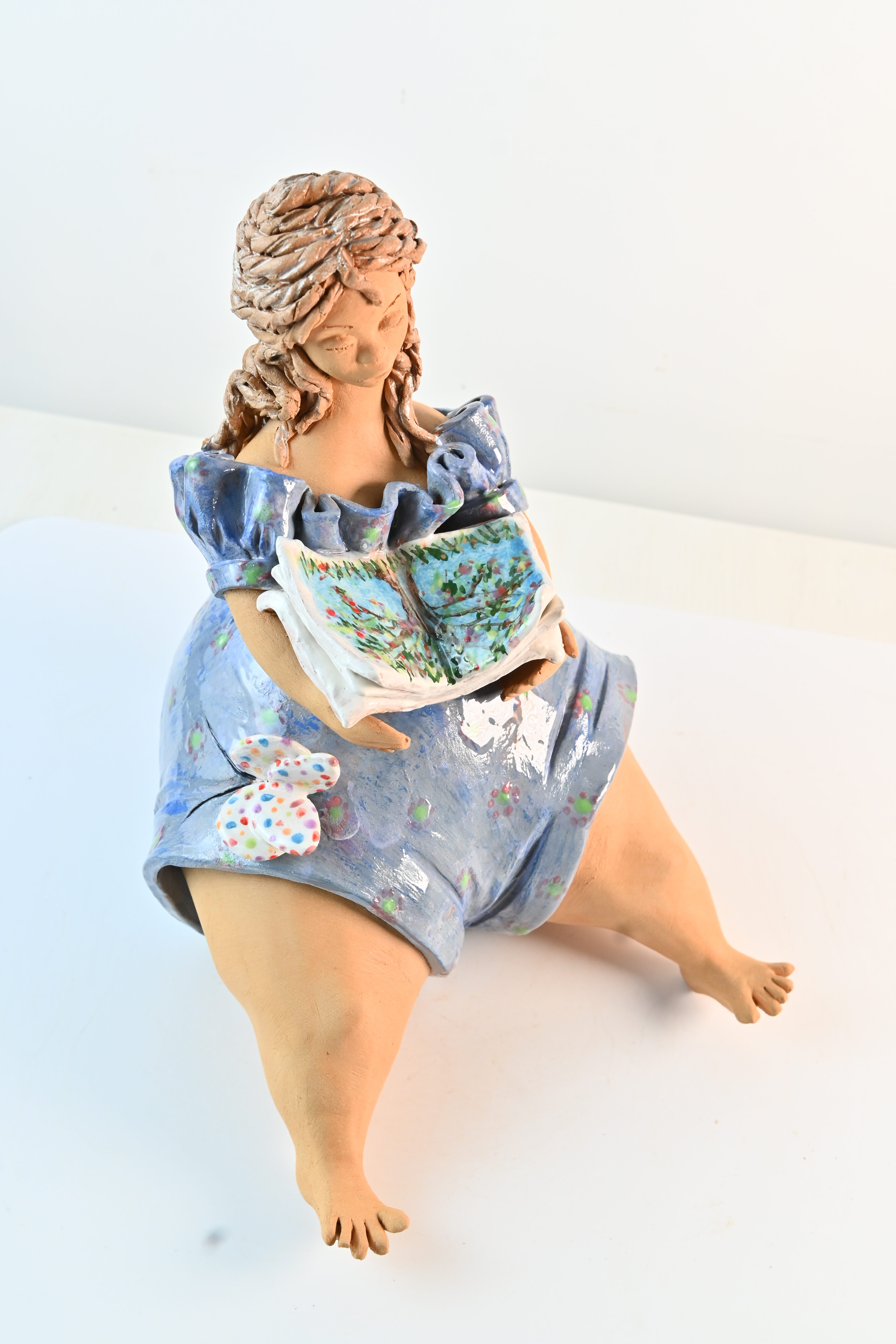 Scultura lettrice con libro seduta con abito azzurro, piedi nudi e farfalla colorata